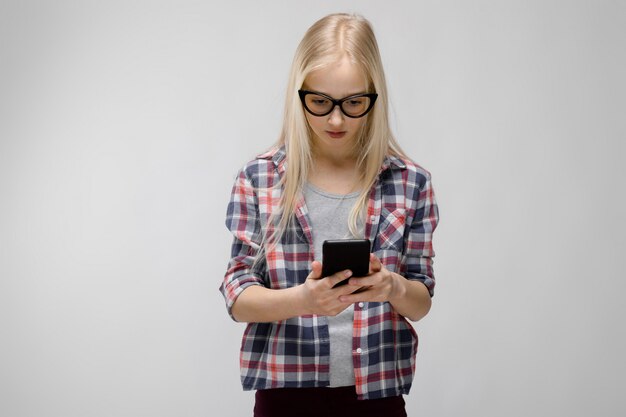 Portrait of attractive smiling sweet adorable blonde teenager girl in checkered clothes in glasses holding mobile phone