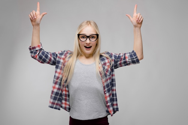 Portrait of attractive smiling sweet adorable blonde teenager girl in checkered clothes in glasses with outstretched hands showing up  gray  