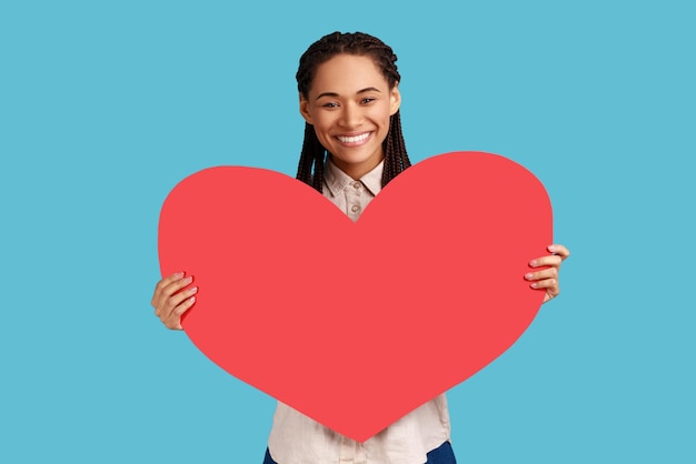 Photo portrait of attractive smiling woman with black dreadlocks holding big red heart expressing positive romantic emotions wearing white shirt indoor studio shot isolated on blue background