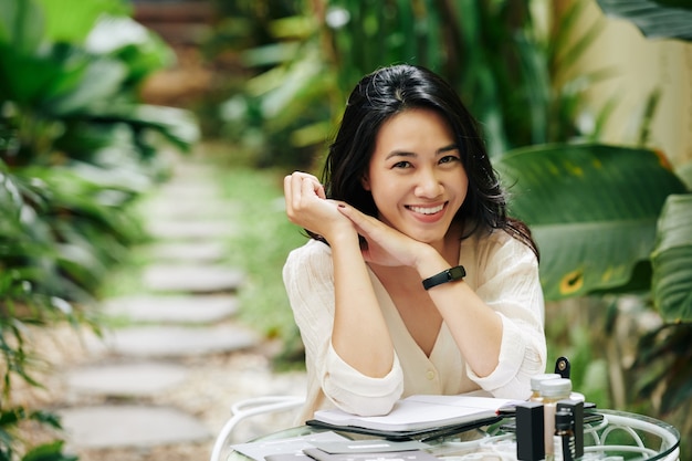 Portrait of attractive smiling young Asian woman sitting at table in backyard and working on business plan for her cosmetics brand