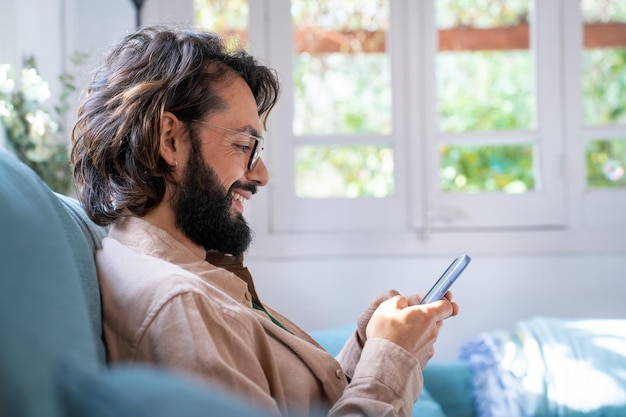 Портрет привлекательного улыбающегося молодого бородатого мужчины, сидящего на диване с помощью смартфона
