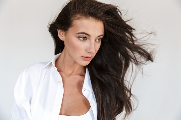 Portrait of an attractive smiling young brunette woman wearing white shirt standing isolated, posing