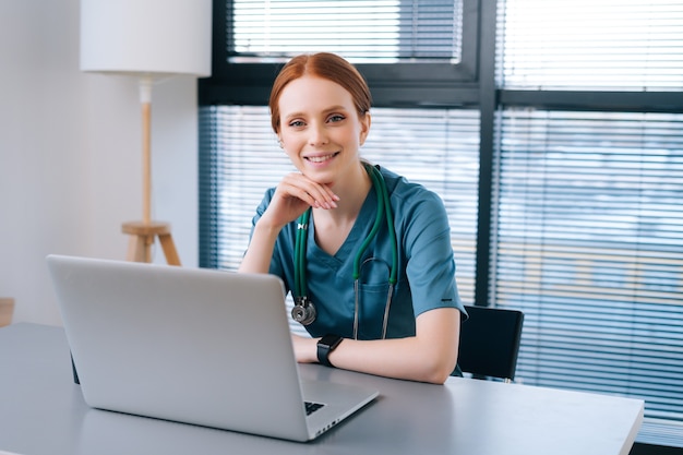 Portrait of attractive smiling young female doctor in blue green medical uniform sitting at desk with laptop on background of window