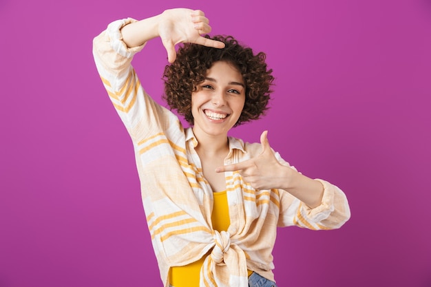 Portrait of an attractive smiling young woman with curly brunette hair standing isolated over violet wall, making photo camera gesture