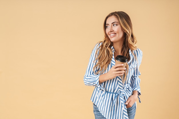 Ritratto di un'attraente giovane donna sorridente con lunghi capelli biondi ricci che indossa abiti casual in piedi isolato sul muro beige, tenendo una tazza di caffè da asporto, guardando lontano