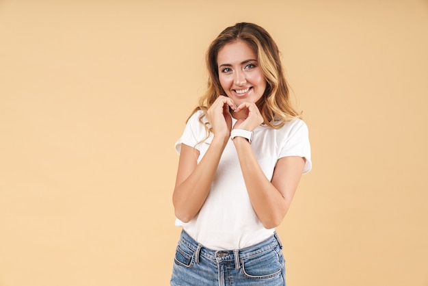 Portrait of an attractive smiling young woman with long curly blonde hair wearing casual clothing standing isolated, showing heart gesture