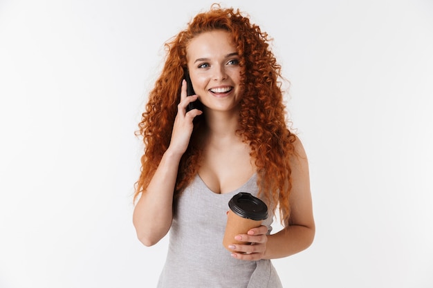 Portrait of an attractive smiling young woman with long curly red hair standing isolated, talking on mobile phone while drinking takeaway coffee