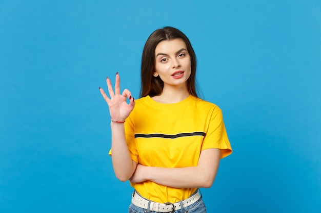 Portrait of attractive stunning young woman in vivid casual clothes looking camera, showing OK gesture isolated on bright blue wall background in studio. People lifestyle concept. Mock up copy space.