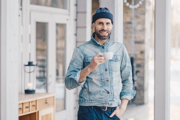 Portrait of attractive stylish bearded male with happy look wears fashionable denim jacket hat and trousers holds mug of hot beverage recreats at home after hard working day smiles pleasantly