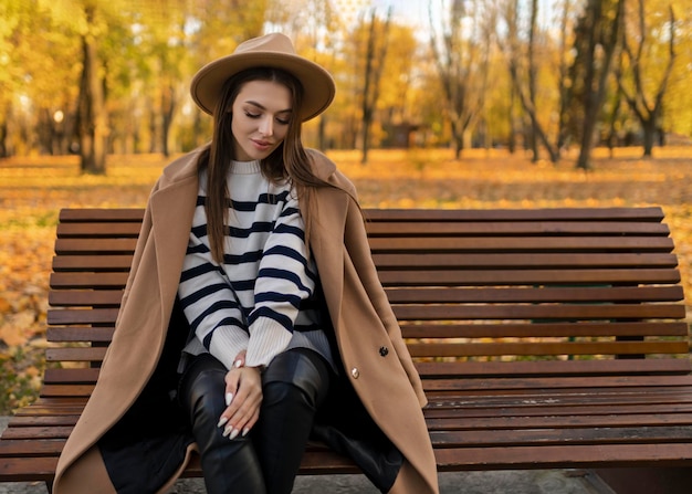 Портрет привлекательной стильной улыбающейся женщины с длинными волосами, прогуливающейся по парку, одетой в теплое коричневое пальто, осенний модный уличный стиль в шляпе