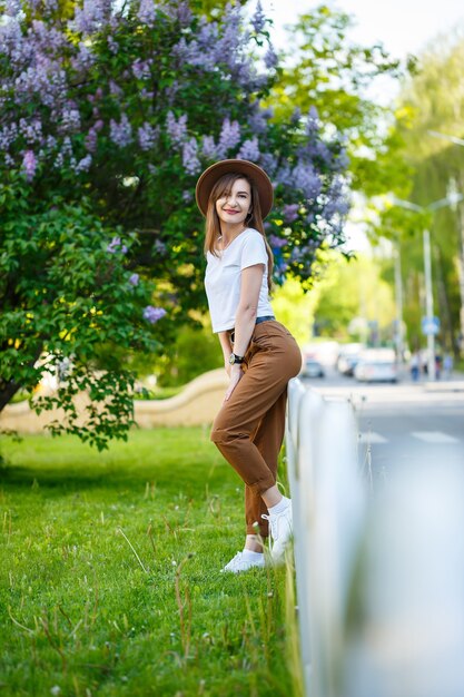 Portrait of attractive stylish woman in a hat with long blonde hair in the park, dressed in a white t-shirt and brown trousers.