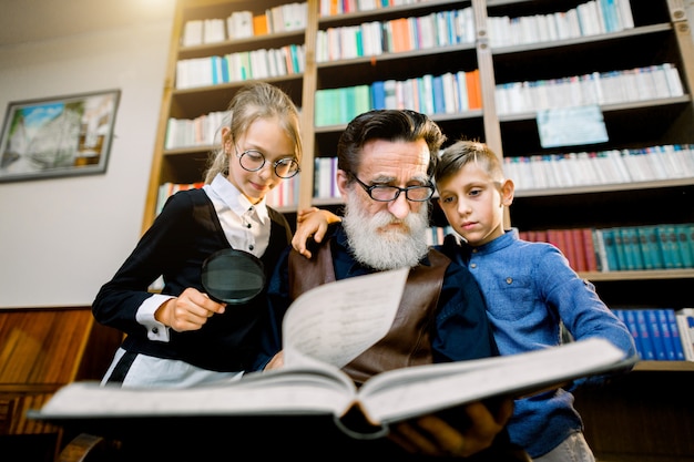 portrait of attractive teen grandchildren and experienced senior bearded grandfather which spending time together in the library on reading interesting book. Bookcase on the background