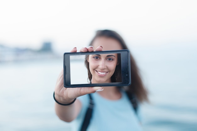 Портрет привлекательной девушки-подростка, стоящей на летнем песчаном пляже в отпуске, держа в руках смартфон, фотографирующий себя в отпуске на фоне голубого неба. Люди путешествуют по технике.