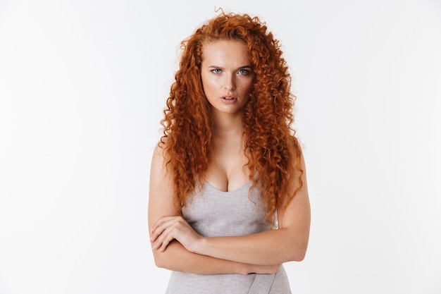 Portrait of an attractive upset young woman with long curly red hair standing isolated, arms folded