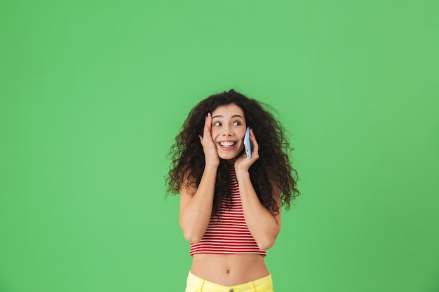 Portrait of attractive woman 20s wearing summer clothes smiling and talking on cell phone isolated on green