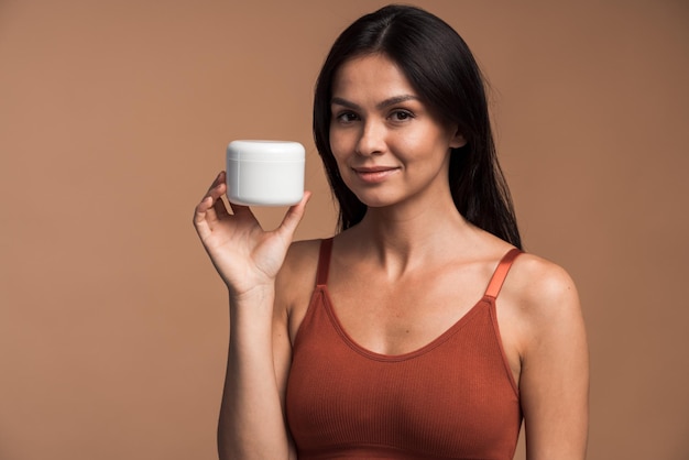 Portrait of attractive woman holding container with face cream and positively looking at camera, isolated on beige color background