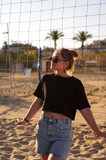 Portrait of attractive woman near volleyball net on the beach.\
beautiful young woman in shorts, black tank top and sunglasses.\
against the background of the beach and palms. vertical photo