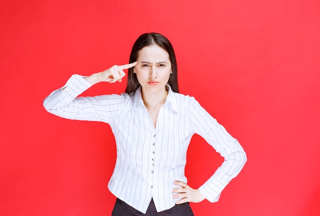 Portrait of attractive woman standing and pointing finger at temple against red wall. 