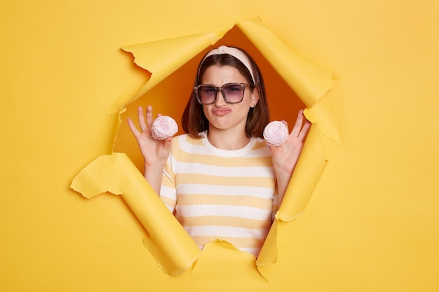 Portrait of attractive woman wearing hair band and striped t shirt standing in yellow paper hole holding marshmallow and frowning face being on diet looking at camera