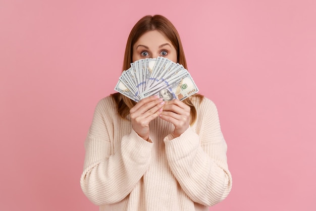 Portrait of attractive young adult blond woman covering half of face with many dollar banknotes, looking at camera, wearing white sweater. Indoor studio shot isolated on pink background.