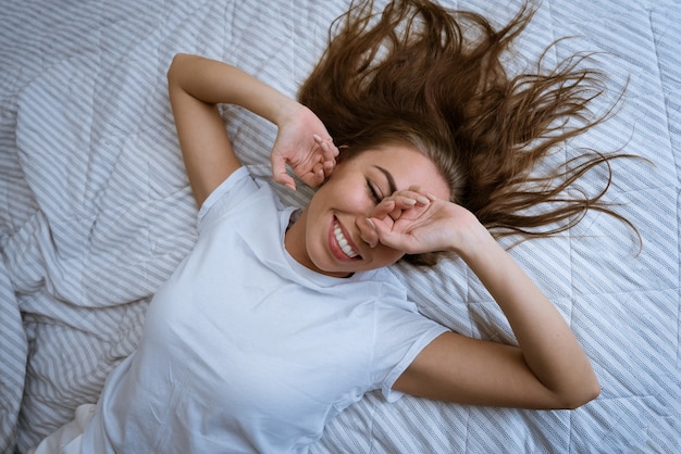 乱れたhaとベッドに横たわっている白いTシャツの魅力的な若い大人のブロンドの女性の肖像画...