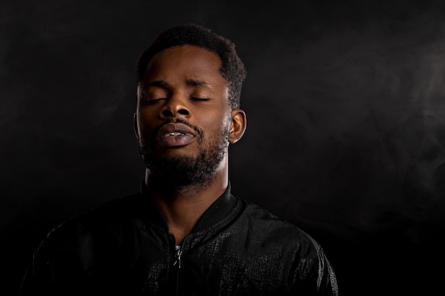 Portrait of attractive young afro man with small beard wearing black jacket looking away withwith closed eyes and thoughtful expression on dark background. Human emotions and facial expressions.