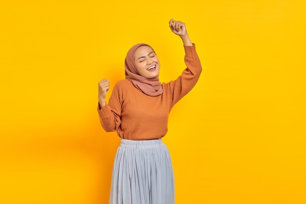 Portrait of attractive young Asian woman raises arms and dances to favorite music isolated on yellow background