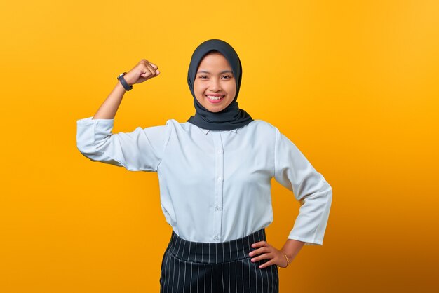 Portrait of attractive young Asian woman showing muscle on yellow background