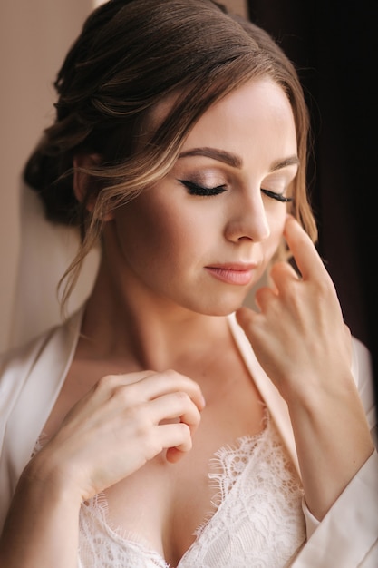 Portrait of attractive young bride in negligee. Beautiful woman stand by window