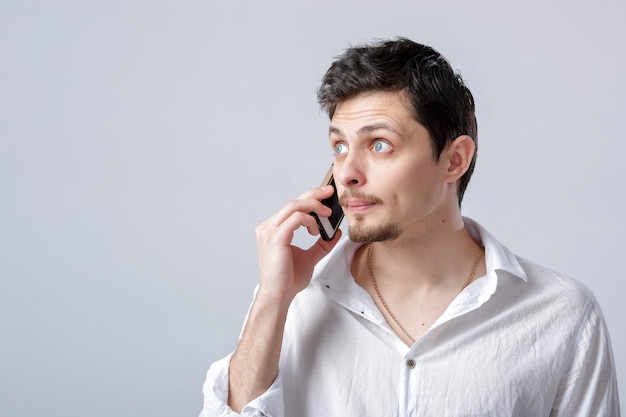 Portrait of attractive young brunette man in white shirt talking on the smartphone on the grey background.