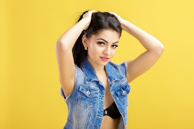 Portrait of attractive young brunette woman in a denim vest on a yellow.