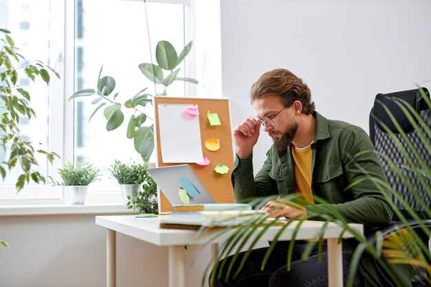 portrait of attractive young businessman using laptop computer at office desk. paperwork and other items. caucasian european bearded guy in casual wear outfit is concentrated on work thinking