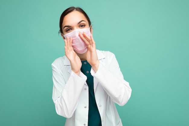 Portrait of an attractive young female doctor in white coat and medical mask standing isolated on blue background