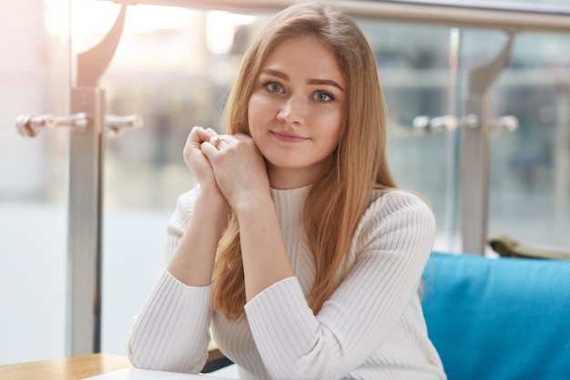 Portrait of attractive young girl being in good mood, has rest in cafeteria after lectures in university, sits on comfortable blue sofa, wears casually, looks at camera, keeps hands folded under chin.