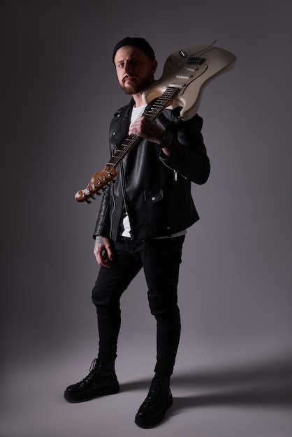 Portrait of an attractive young guy in a leather jacket and tattoos with an electric guitar on his shoulder Dark background