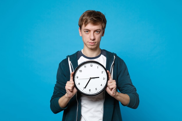 Portrait of attractive young man in casual clothes holding round clock isolated on blue wall . Time is running out. People sincere emotions, lifestyle concept. 