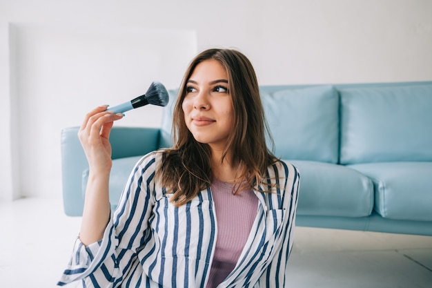 Portrait of attractive young woman holding makeup brush in hand sitting on floor near blue sofa