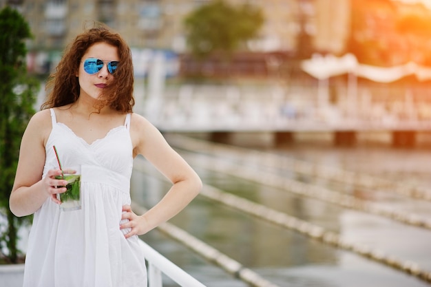 Ritratto di una giovane donna attraente in posa con il suo cocktail su una riva del lago che indossa occhiali da sole