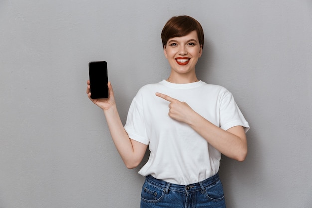 Portrait of attractive young woman smiling and pointing finger at cellphone isolated over gray wall