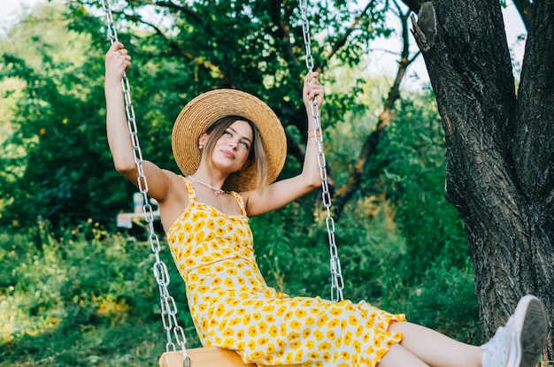 Portrait of attractive young woman in straw hat outdoors in summer sunny day on a swing.