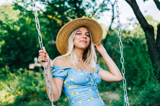 ブランコに乗って夏の晴れた日に屋外で麦わら帽子の魅力的な若い女性の肖像画。