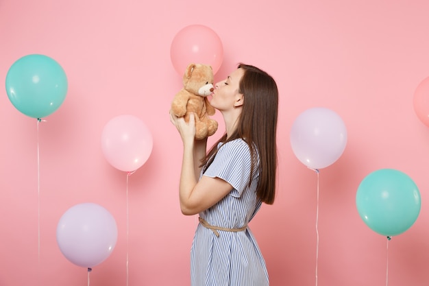 Portrait of attractive young woman wearing blue dress holding and kissing teddy bear plush toy on pink background with colorful air balloons. Birthday holiday party, people sincere emotions concept.
