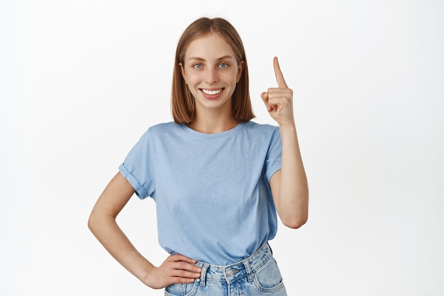 Portrait of attractive young woman with blond hair and blue eyes, pointing finger up at promo, showing sale dicount banner and smiling satisfied, white wall.