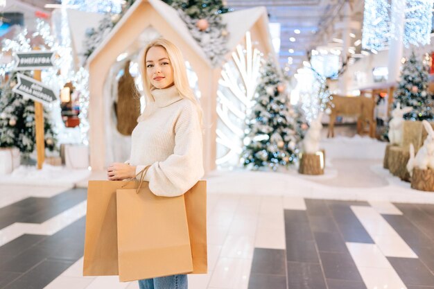 Portrait of attractive young woman with blonde hair holding bags with purchase looking at camera standing in hall of celebrate shopping mall in Christmas eve