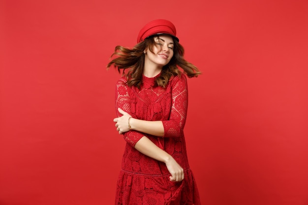 Portrait of attractive young woman with flowing hair in lace dress and cap holding hands crossed isolated on bright red wall background. People sincere emotions, lifestyle concept. Mock up copy space.