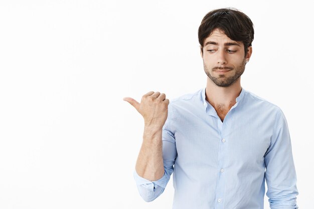 Portrait of awkward ashamed and intense good-looking young man with beard hiding eyes as looking aside pointing left embarrassed over gray wall