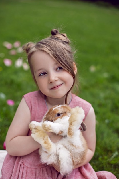 Ritratto bambina in un abito rosa gioca con un coniglio in un prato verde in estate divertente amicizia tra un bambino e un animale durante la pasqua