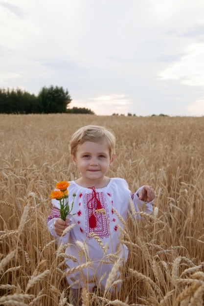 Portrait of baby in shirt with red embroidery and bouquet of flowers in her hand in wheat field at sunset