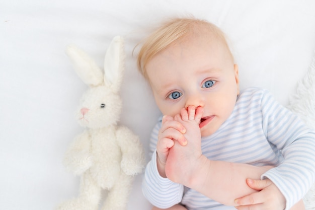 Portrait of baby sucking foot lying on bed, baby boy blonde six months