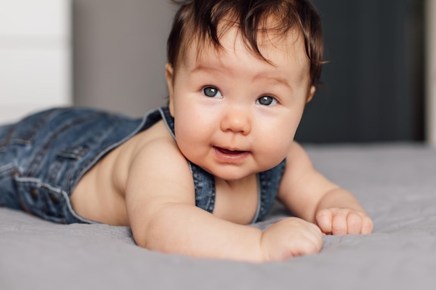 自宅で灰色の毛布の上に横たわっているデニムロンパースを着ている赤ちゃんの肖像寝室でベッドの上を這う青い目の幼児の子供を見上げる